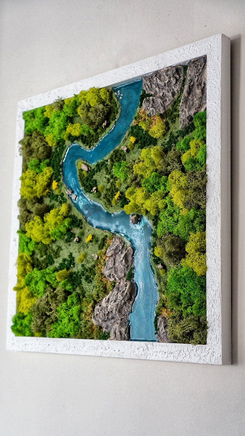 Puede incluir: Una pieza de arte de musgo enmarcada que representa un r&iacute;o serpenteando a trav&eacute;s de un exuberante bosque verde. El r&iacute;o es de un azul vibrante y el musgo es de varios tonos de verde.
