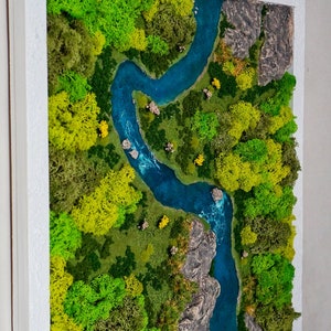 Puede incluir: Una pieza de arte de musgo enmarcada que representa un r&iacute;o serpenteando a trav&eacute;s de un bosque verde. El r&iacute;o es de un azul vibrante y el bosque est&aacute; hecho de musgo preservado en tonos de verde y amarillo.