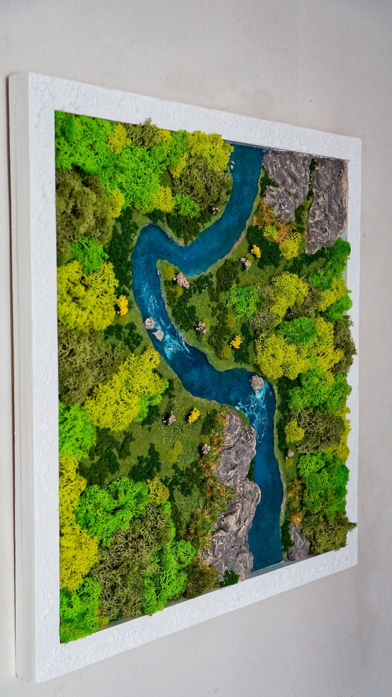 Puede incluir: Una pieza de arte de musgo enmarcada que representa un r&iacute;o serpenteando a trav&eacute;s de un bosque verde. El r&iacute;o es de un azul vibrante y el bosque est&aacute; hecho de musgo preservado en tonos de verde y amarillo.