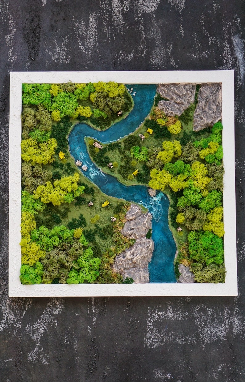 Puede incluir: Una pieza de arte de musgo enmarcada que representa un r&iacute;o serpenteando a trav&eacute;s de un bosque verde. El r&iacute;o es de un azul vibrante y el bosque est&aacute; hecho de musgo preservado en tonos verdes y amarillos.