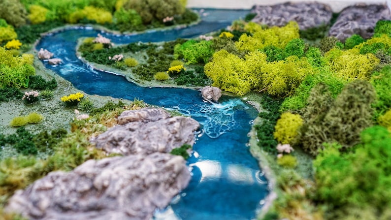 Puede incluir: Un paisaje en miniatura con un r&iacute;o de resina azul serpenteando a trav&eacute;s de musgo verde y rocas grises.