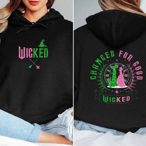 May include: Black hoodie featuring the word "Wicked" in pink and green glitter, with a witch hat and crossed brooms. The back displays a design with the words "Changed for Good" and the word "Wicked".