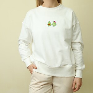 Könnte beinhalten: Weißer Pullover mit einem niedlichen gestickten Design aus zwei Avocados, die sich an den Händen halten. Die Avocado rechts hat eine rosa Schleife.