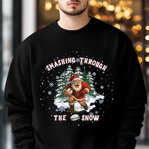 Puede incluir: Sudadera negra con un diseño gráfico de Papá Noel jugando al rugby en un paisaje nevado. El texto "SMASHING THROUGH THE SNOW" está presente. También se incluyen copos de nieve y pinos en el diseño.