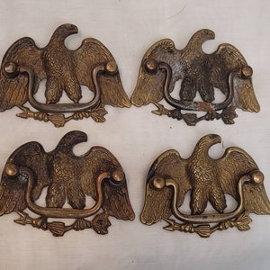 Pode incluir: Quatro puxadores de gaveta antigos em latão em forma de águia. Cada puxador apresenta uma águia detalhada com asas abertas, uma alça curva e um design de flecha e folha. Os puxadores têm uma aparência vintage e envelhecida.