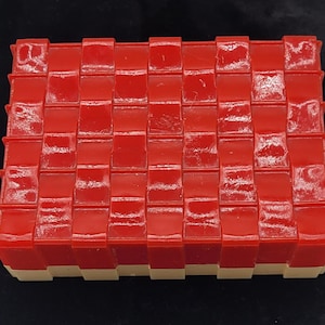 May include: A rectangular object with a woven pattern, featuring a bright red top and a cream-coloured base. The design consists of repeating rectangular blocks, creating a textured surface. The object appears to be made of a glossy material.