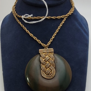 Könnte beinhalten: Eine goldene Kettenkette mit einem großen, runden Anhänger in Dunkelgrün und Braun. Der Anhänger hat ein geflochtenes Golddesign in der Mitte. Das Etikett an der Kette lautet "Max Factor Thalal Cachet Perfume."