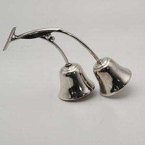 May include: A silver double bell with a decorative handle. The bells are attached to a curved stem and have a smooth, polished finish.