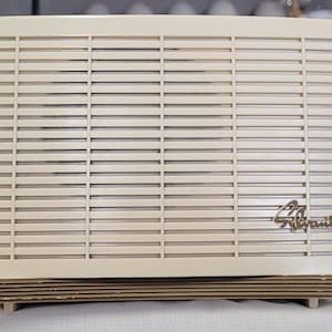 Op de afbeelding: Crèmekleurige vintage Sylvania elektrische kachel met horizontale lamellen en een temperatuurknop. Het Sylvania-logo is zichtbaar op de voorkant. De kachel heeft een rechthoekige vorm en een retro-ontwerp, wat suggereert dat hij uit het midden van de 20e eeuw komt.