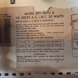 May include: Close-up of a vintage radio's back panel with instructions for using 115 volts AC/DC or battery power. The panel is beige with black text and a diagram showing battery placement. The text includes model number 879 Series B, 20 watts, and battery types Eveready #717 and #467.