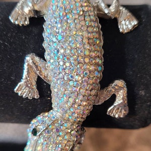 Op de afbeelding: Een zilveren hagedis broche bedekt met iriserende strass steentjes. De hagedis heeft een groen oog en staat in een looppositie.