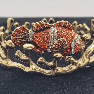 May include: A gold-tone bracelet with a detailed design of coral and a red and white jeweled fish. The fish is set with red and clear rhinestones.