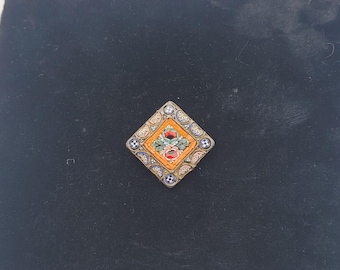 Puede incluir: Un broche de mosaico de forma cuadrada con un diseño floral en tonos naranja, rojo, verde y azul. El broche está engastado en un marco de metal plateado.