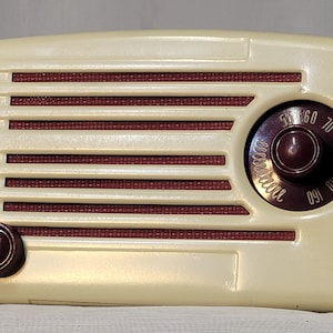 Puede incluir: Una radio vintage blanca y marrón con un dial redondo y una perilla en el lateral. El dial tiene números del 55 al 160.