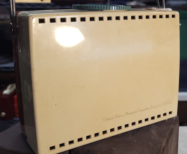 May include: A vintage cream-colored Emerson Radio and Phonograph Corporation, Jersey City, N.J. USA radio with a green knob and a metal grill.
