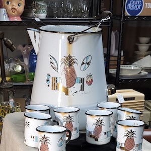May include: A set of eight white enamel mugs with black trim and a pineapple design. The mugs are arranged in a circle on a white tablecloth. A large white enamel pitcher with a pineapple design is in the background.
