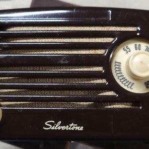 Puede incluir: Una radio Silvertone vintage de color marrón con un dial y botones blancos. La radio tiene una rejilla metálica y una etiqueta blanca con el nombre de la marca.