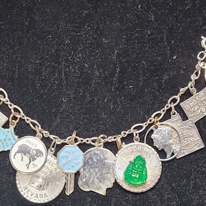 May include: Silver charm bracelet featuring various charms, including state shapes, a coin, a key, and a green jade Buddha. The charms are connected by a silver chain. Some charms have text, such as "KENTUCKY" and "NEVADA".