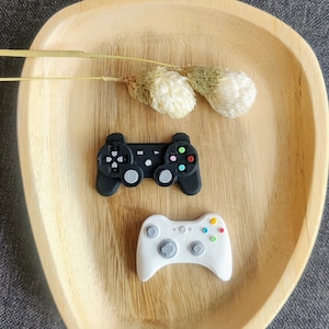 Könnte beinhalten: Zwei Miniatur-Videospielcontroller aus Plastik, einer schwarz und einer weiß, werden auf einem Holztablett präsentiert. Das Tablett ist oval geformt und hat eine natürliche Holzoptik.