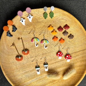 Può includere: Una collezione di orecchini in argilla polimerica a tema autunnale, tra cui zucche, funghi, tazze da caffè, foglie e ombrelli. Gli orecchini sono esposti su un piatto di legno.