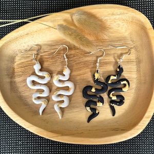 Könnte beinhalten: Ein Paar weiße und schwarze Schlangenohrringe mit goldenen Akzenten. Die Ohrringe sind aus Polymer Clay gefertigt und haben ein baumelndes Design.