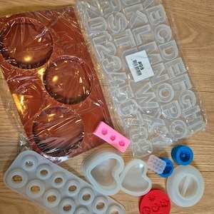 Può includere: Una collezione di stampi in silicone che include un vassoio rettangolare marrone con tre cavità circolari, un foglio trasparente con lettere e numeri, uno stampo ad anello bianco, due stampi a forma di cuore, un piccolo stampo a rosa rosa e vari piccoli stampi circolari.