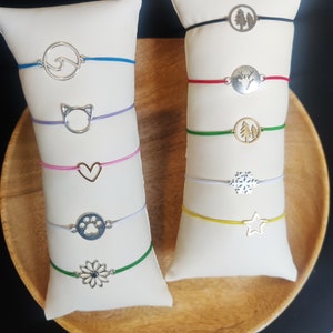 May include: Two white display stands with colorful string bracelets featuring various silver charms. The charms include a wave, cat, heart, paw print, flower, trees, deer, snowflake, and star.