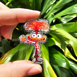 Puede incluir: Un personaje rojo y naranja con purpurina, con una gran cabeza roja y blanca a rayas y un cuerpo rojo. El personaje está saludando con la mano derecha y tiene una gran sonrisa.