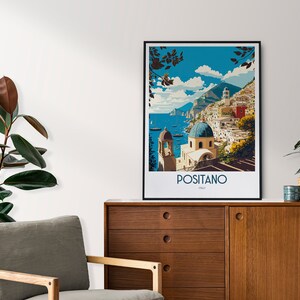 May include: A vintage-style travel poster featuring a colorful illustration of Positano, Italy. The poster depicts a seaside town with buildings on a hillside, a blue sky with clouds, and a blue ocean with boats. The text "Positano, Italy" is printed at the bottom of the poster.