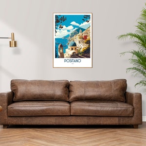May include: A brown leather sofa with two cushions in front of a white wall with a framed poster of Positano, Italy. The poster features a colorful illustration of the town with buildings, a blue sea, and a mountain in the background. A potted plant with green leaves is on the right side of the image.