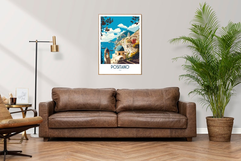 May include: A brown leather sofa with two cushions in front of a white wall with a framed poster of Positano, Italy. The poster features a colorful illustration of the town with buildings, a blue sea, and a mountain in the background. A potted plant with green leaves is on the right side of the image.