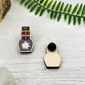 Peut inclure: Épinglette en bois en forme de vase, ornée d'une fleur blanche et de motifs floraux colorés. L'épingle a un fermoir noir au dos. Le design s'inspire de l'art populaire traditionnel.