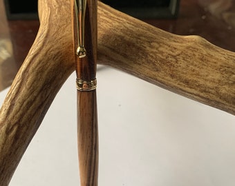 May include: A wooden pen with a gold-colored top and bottom. The pen is resting on a piece of wood with a brown and white striped pattern.