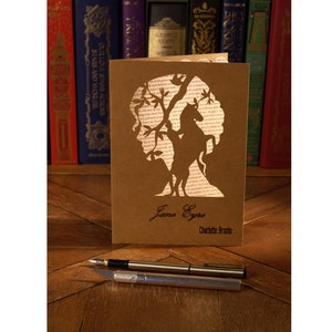 May include: A tan paper greeting card featuring a die-cut silhouette of a person and a horse under a tree, with the text Jane Eyre and Charlotte Bronte printed below. A silver fountain pen and a silver craft knife are positioned in front of the card.