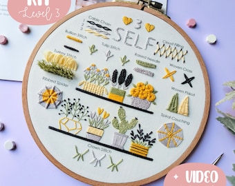 May include: A white embroidery hoop with a beige fabric inside. The fabric has various embroidery stitches and designs, including a cable chain, bullion stitch, tulip stitch, ribbed spider, crown stitch, raised needle, woven cross, woven piskot, spiral couching, and cloud filling. The hoop has the text "KIT Level 3" and "SELF" on it.