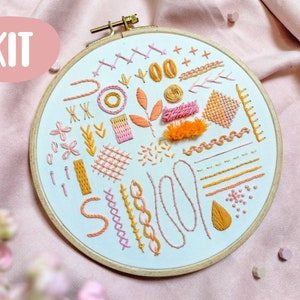 Op de afbeelding: Borduurpakket met een houten ring met verschillende gestikte patronen in oranje, roze en geel. Het woord "KIT" staat in een roze ovaal. Het ontwerp bevat bloemen en geometrische elementen. De ring heeft een diameter van ongeveer 20 cm.