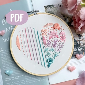 Puede incluir: Diseño de corazón bordado en un aro de madera. El corazón está lleno de bordados florales y lineales coloridos en tonos rosa, turquesa y melocotón. La palabra "PDF" es visible en un círculo lila claro.