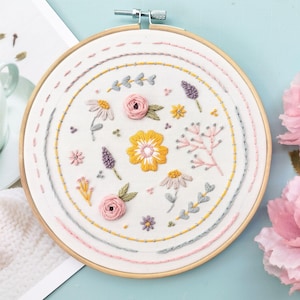 Pu&ograve; includere: Un telaio da ricamo circolare in legno contenente tessuto bianco con disegni floreali cuciti a mano. Il ricamo presenta un fiore giallo centrale circondato da rose rosa, piccoli rametti viola e vari delicati motivi botanici nelle tonalit&agrave; del giallo, rosa e grigio, incorniciati da cerchi cuciti concentrici.