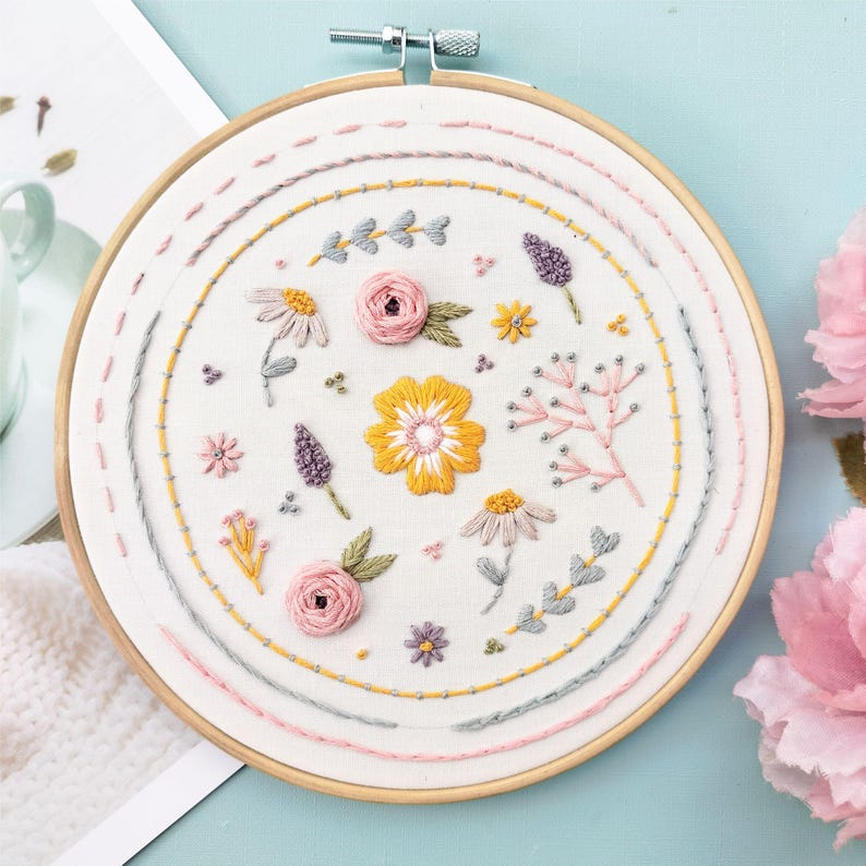 Pu&ograve; includere: Un telaio da ricamo circolare in legno contenente tessuto bianco con disegni floreali cuciti a mano. Il ricamo presenta un fiore giallo centrale circondato da rose rosa, piccoli rametti viola e vari delicati motivi botanici nelle tonalit&agrave; del giallo, rosa e grigio, incorniciati da cerchi cuciti concentrici.