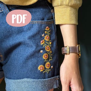 Puede incluir: Shorts vaqueros azules con dobladillo vuelto y bolsillo. Los shorts presentan un diseño bordado de girasoles y abejas. Un círculo rosa con la palabra "PDF" está en la esquina superior izquierda.