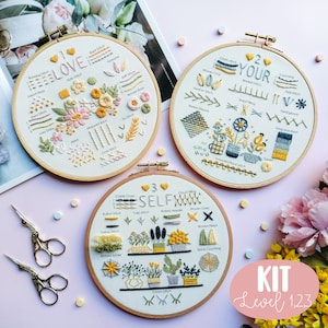 Könnte beinhalten: Drei Stickrahmen mit Blumenmustern und Stich-Anleitungen. Die Rahmen sind mit "I Love", "Your" und "Self" mit Herzen und Zahlen beschriftet. Die Rahmen befinden sich auf einer rosa Oberfläche mit einer rosa und gelben Blume im Hintergrund. Der Text "KIT Level 1,2,3" befindet sich in einem rosa Kreis.