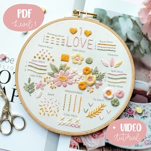 Può includere: Un cerchio da ricamo bianco con un motivo floreale e la parola "LOVE" ricamata in filo rosa e giallo. Il cerchio presenta una varietà di punti di ricamo, tra cui il punto a nodo, il nodo francese, il punto a raso, il punto indietro, il punto a spina di pesce, il punto a gambo, la margherita pigra, il punto diviso, la rosa tessuta, il punto a seme, il punto a frusta, il punto a mosca e il punto a croce.