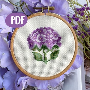Peut inclure: Broderie au point de croix d'une fleur d'hortensia violette avec des feuilles vertes, dans un cercle en bois. Le mot "PDF" est dans un cercle violet en haut à gauche. Le fond comprend des fleurs et de la verdure violettes.