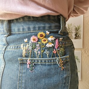 Pu&ograve; includere: Un paio di jeans blu denim con una tasca ricamata con una variet&agrave; di fiori colorati, tra cui rosa, gialli e bianchi.