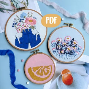 以下が含まれることがあります： 3つの刺繍フープ。1つは青いジーンズと花を持つ手、もう1つは「fuck this」の文字と花、3つ目はピンクの背景に黄色の柑橘系のスライス。木製のフレームに入った、さまざまなデザインの刺繍フープ。
