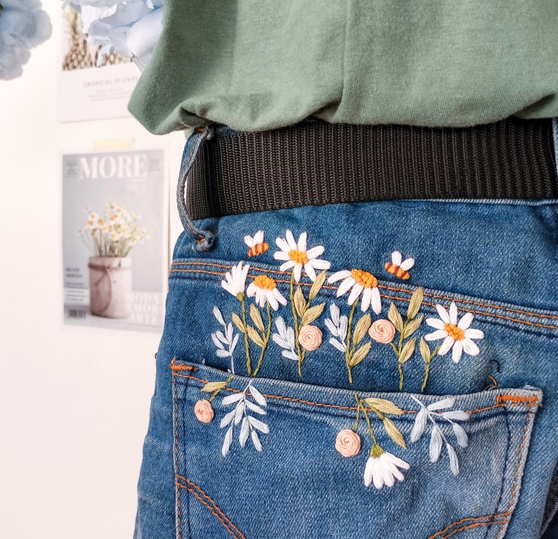 Peut inclure: Un jean bleu denim avec une poche arri&egrave;re brod&eacute;e de marguerites blanches et de feuilles vertes.
