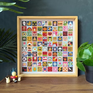 Puede incluir: Una obra de arte de punto de cruz enmarcada con una cuadrícula de imágenes pixeladas coloridas. Las imágenes incluyen un pato, una hamburguesa, un corazón y la palabra «OK». El marco es de madera clara y la obra se exhibe sobre una superficie de madera.