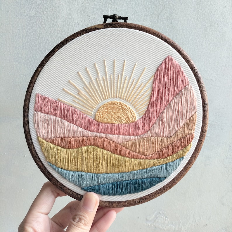 May include: Embroidered hoop art featuring a sunset over a landscape with layered hills in shades of pink, orange, yellow, and blue.