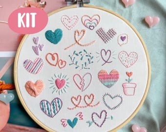 May include: A round embroidery hoop with a variety of embroidered hearts in different colors and styles. The word "KIT" is in a pink oval in the upper left corner. Pink heart-shaped embellishments are scattered around the hoop.