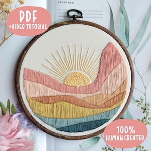 May include: A circular wooden embroidery hoop containing a fabric panel with a stitched landscape design. The embroidery features a yellow sun with radiating rays above layered hills in shades of pink, peach, mustard yellow, and teal green.